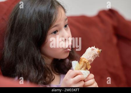 La jeune fille expressive mange du poulet et des légumes pour le dîner sur le canapé tout en regardant la télévision Banque D'Images