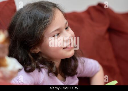 La jeune fille expressive mange du poulet et des légumes pour le dîner sur le canapé tout en regardant la télévision Banque D'Images