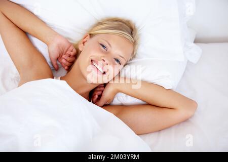 Profiter des effets d'une bonne nuit de repos. Vue en grand angle d'une jeune femme allongé au lit et souriant à la caméra. Banque D'Images