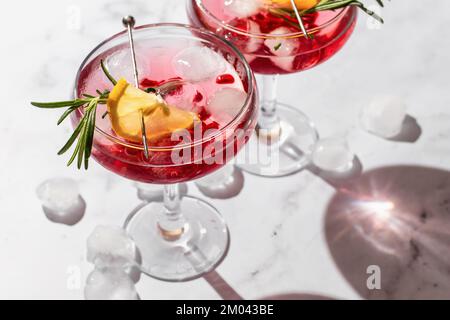 Deux verres de cocktail glacé à la grenade avec romarin et citron sur fond de marbre blanc, isolés avec des ombres claires dans des conditions de forte lumière. Noël Banque D'Images