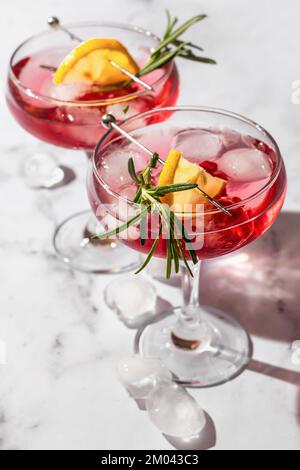 Deux verres de cocktail glacé à la grenade avec romarin et citron sur fond de marbre blanc, isolés avec des ombres claires dans des conditions de forte lumière. Noël Banque D'Images