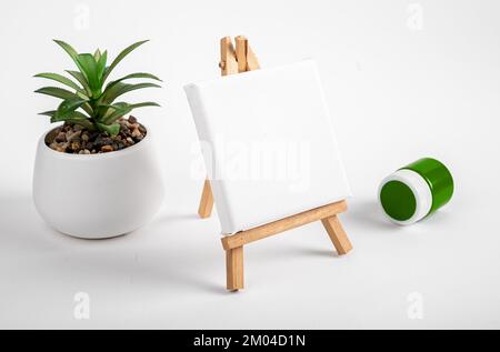 Maquette de toile blanche vierge en angle de perspective, mini trépied en bois, chevalet et plante de maison en pot, peinture verte dans un pot sur le bureau. Photo de haute qualité Banque D'Images