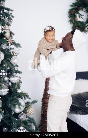 L'homme africain heureux a du plaisir et joue avec sa fille de race mixte sur fond d'arbre de Noël. Le concept de la famille interraciale et de l'unité soit Banque D'Images