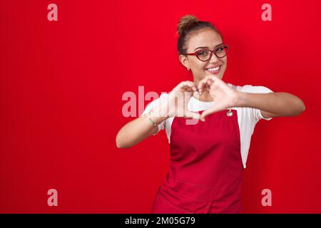 Jeune femme hispanique portant un tablier de serveuse sur fond rouge souriant dans l'amour faisant le symbole de forme de coeur avec les mains. concept romantique. Banque D'Images