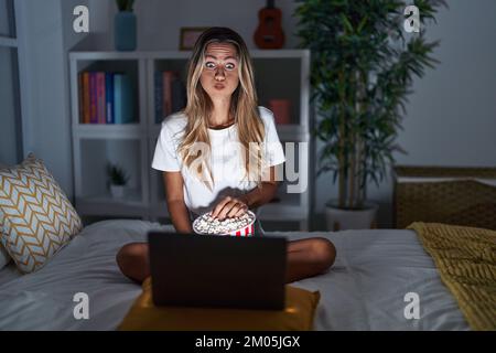 Jeune femme blonde assise sur le lit à la maison regardant un film d'ordinateur portable puffant des joues avec le visage drôle. bouche gonflée avec de l'air, attrapant l'air. Banque D'Images