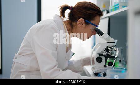 Femme hispanique d'âge moyen portant l'uniforme scientifique au microscope en laboratoire Banque D'Images