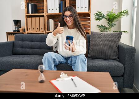 Jeune femme hispanique allant à la thérapie au bureau de consultation souriant heureux et positif, pouce vers le haut faisant excellent et signe d'approbation Banque D'Images