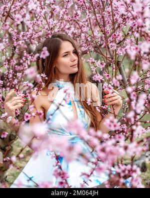 Belle fille souriante assise sur un arbre de pêche en fleur dans une robe blanc-bleu, cheveux longs. Bonne humeur. Heure de printemps. Banque D'Images