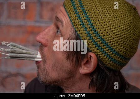 Homme poilu avec une casquette verte et dix joints de marijuana près de vieux mur rouge de brique Banque D'Images