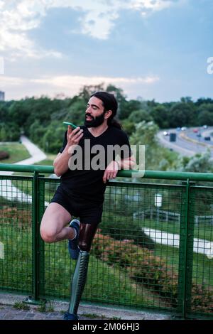 Jeune homme avec une jambe prothétique appréciant un après-midi à l'extérieur tout en envoyant un message audio Banque D'Images