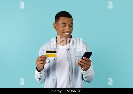 Souriant jeune afro-américain dans la recherche décontractée de smartphone, utilise la carte de crédit pour faire des achats en ligne Banque D'Images