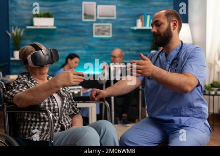 Une femme âgée utilise un casque de réalité virtuelle sur un fauteuil roulant dans une maison de soins pour explorer la réalité augmentée. Infirmière de sexe masculin dans la salle commune de l'établissement de soins qualifiés aidant le patient à s'amuser. Banque D'Images