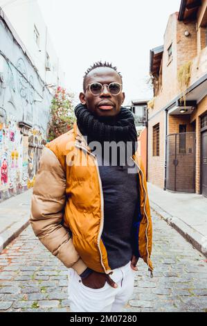 Portrait du jeune Africain, portant des lunettes et des vêtements de mode d'hiver, debout au milieu de la rue, les mains dans ses poches de pantalon a Banque D'Images