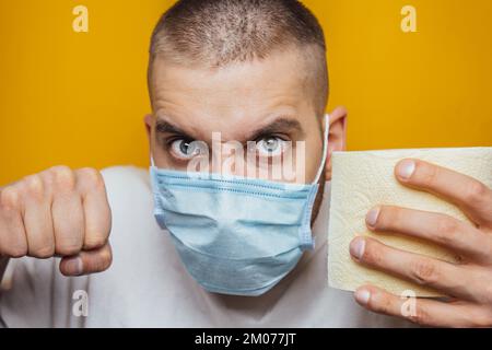Un jeune homme dans un masque médical tient des rouleaux de papier toilette, montre un geste dangereux sur un fond jaune. Le concept de stock, les articles manquants, la panique et le quaran Banque D'Images