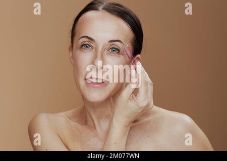 Portrait d'une femme moderne d'âge moyen avec une pince à épiler isolée sur le beige. Banque D'Images