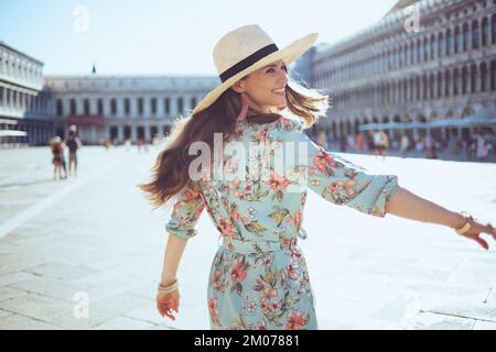 Heureuse voyageur à la mode femme en robe florale avec chapeau ayant excursion à la Piazza San Marco à Venise, Italie. Banque D'Images