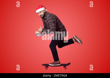 Père Noël à bord d'un skateboard ou d'une longue planche et tenant deux boîtes cadeaux de cadeaux sur fond rouge. Concept de livraison rapide. Arrière-plan Noël Banque D'Images