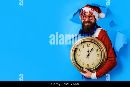 Homme barbu dans le chapeau de père Noël et lunettes de fête avec horloge regardant à travers trou de papier. Ventes de Noël. Banque D'Images