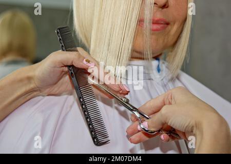 Gros plan d'un coiffeur coupe les cheveux blancs mouillés d'un client dans un salon. Coiffeur coupe une femme. Vue latérale d'un coupe de cheveux à la main avec des ciseaux Banque D'Images