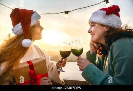 Des amis heureux fêtent Noël en plein air. Deux jeunes femmes s'amusent, boivent du vin, portent des chapeaux de père Noël. Banque D'Images