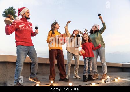 Des amis heureux fêtent Noël en plein air. Les adultes et les enfants s'amusent en portant des chapeaux de père Noël. Banque D'Images