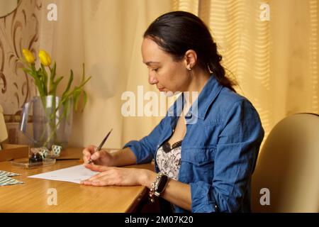 Une fille indienne écrit avec un stylo à encre sur une feuille de papier blanche. papeterie sur un bureau en bois. leçons d'orthographe et exercices de calligraphie. la paperasserie dans l'offic Banque D'Images