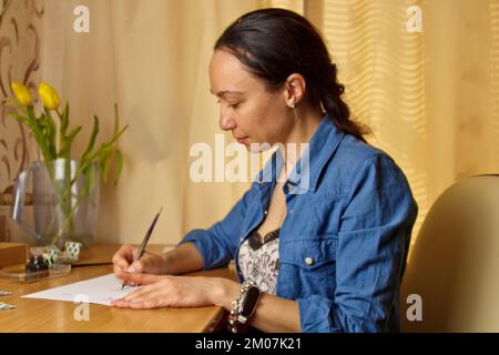 Une fille indienne écrit avec un stylo à encre sur une feuille de papier blanche. papeterie sur un bureau en bois. leçons d'orthographe et exercices de calligraphie. la paperasserie dans l'offic Banque D'Images