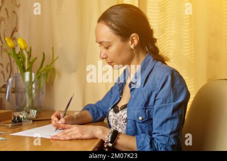 Une fille indienne écrit avec un stylo à encre sur une feuille de papier blanche. papeterie sur un bureau en bois. leçons d'orthographe et exercices de calligraphie. la paperasserie dans l'offic Banque D'Images