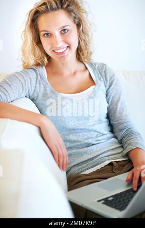 Connecté au monde depuis le confort de la maison. Portrait d'une belle jeune femme qui vous sourit tout en travaillant sur un ordinateur portable à la maison. Banque D'Images