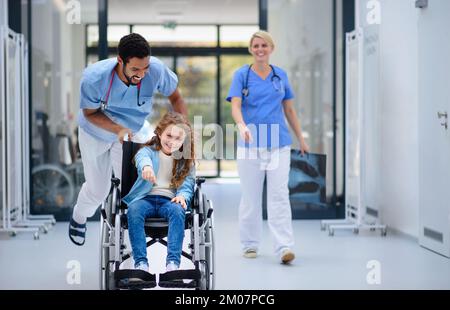 Un jeune médecin pousse une petite fille en fauteuil roulant dans un couloir pédiatrique, s'amusant. Banque D'Images