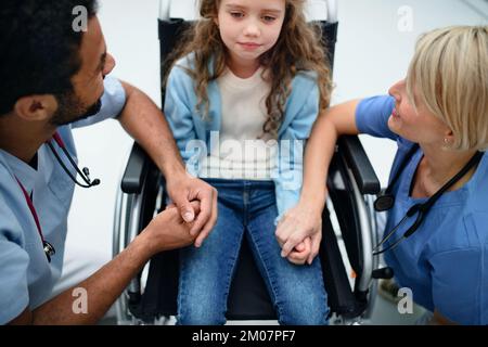 Jeune médecin multiracial avec son collègue consolant une petite fille en fauteuil roulant. Banque D'Images