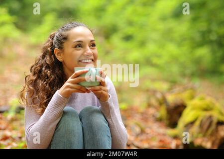 Une femme heureuse assise dans une forêt buvant un café en regardant loin Banque D'Images