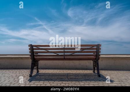 Un banc vide sur fond de ciel bleu et de nuages dans le parc se dresse sur des pavés gris, devant le parapet. Banque D'Images