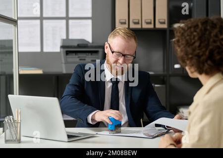 Portrait de l'homme barbu estampant le formulaire de demande tout en travaillant avec une femme visiteur dans le bureau de l'agence Banque D'Images