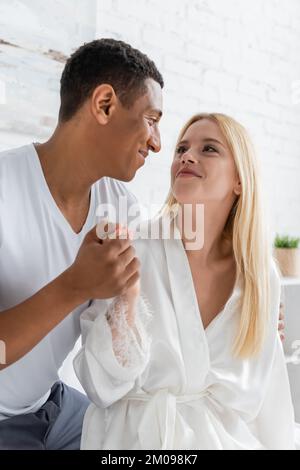 joyeux couple multiethnique en tenues de nuit blanches tenant les mains et regardant l'un l'autre dans la chambre Banque D'Images