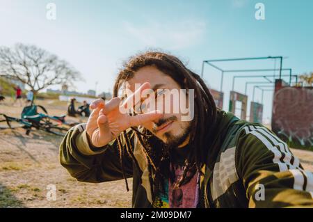 selfie photo avec le téléphone de jeune caucasien vénézuélien avec des dreadlocks regardant l'appareil photo faire la paix et l'amour signe avec la main, v signe avec les doigts, Banque D'Images