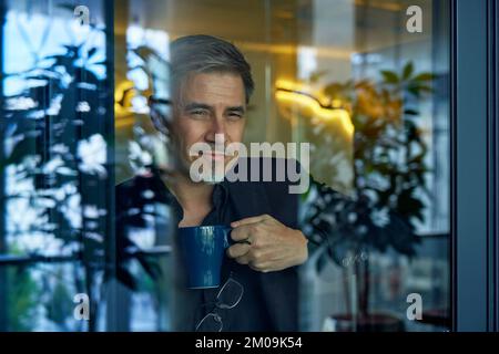 Portrait d'affaires - homme d'affaires regardant par la fenêtre dans le bureau, souriant. Âge mûr, âge moyen, homme adulte moyen en 50s avec un sourire heureux et confiant. Copier l'espace. Banque D'Images