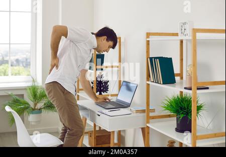 Un homme qui travaille sur un ordinateur portable ressent de la douleur dans son dos, se lève et se moque de son dos pour se détendre. Banque D'Images