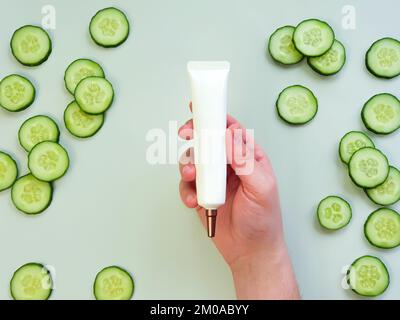 Tube de crème pressée blanc sans marque avec buse longue et bouchon à vis en bronze dans la main d'un homme. Bouteille pour la marque et tranches de concombre frais sur vert clair Banque D'Images