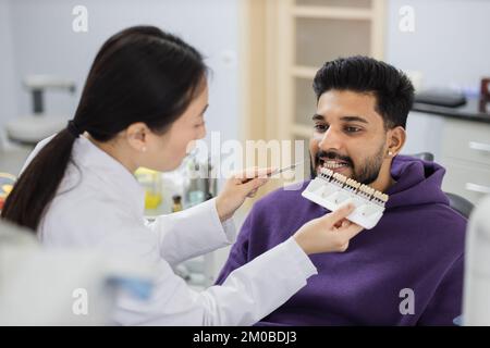 Un dentiste asiatique vérifie et sélectionne la couleur des dents des jeunes hommes barbus. Méthode visuelle de perception subjective couleur de remplissage par un médecin. Dentiste choisissant la couleur de remplissage à partir du concept de palette Banque D'Images