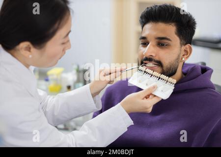 Un dentiste asiatique vérifie et sélectionne la couleur des dents des jeunes hommes barbus. Méthode visuelle de perception subjective couleur de remplissage par un médecin. Dentiste choisissant la couleur de remplissage à partir du concept de palette Banque D'Images