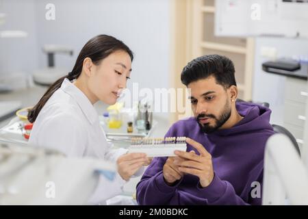 Un dentiste asiatique vérifie et sélectionne la couleur des dents des jeunes hommes barbus. Méthode visuelle de perception subjective couleur de remplissage par un médecin. Dentiste choisissant la couleur de remplissage à partir du concept de palette Banque D'Images