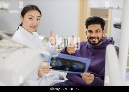 Beau jeune homme barbu, assis sur une chaise de dentiste et regardant la radiographie de l'image scanner des dents avec sa femme joyeuse dentiste asiatique à la clinique montrant le pouce vers le haut. Banque D'Images