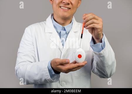 Sourire mature chinois gars thérapeute en manteau blanc montre sos bouton d'alarme, isolé sur fond gris, rogné Banque D'Images