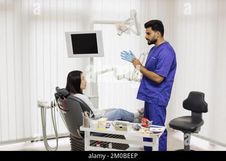 Un dentiste mâle barbu confiant met des gants en caoutchouc bleu, debout dans un cabinet dentaire, prêt pour l'examen et le traitement de sa patiente. Concept de patient et de dentiste heureux Banque D'Images