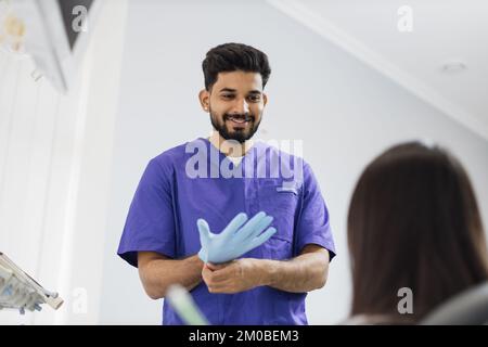 Un dentiste mâle barbu confiant met des gants en caoutchouc bleu, debout dans un cabinet dentaire, prêt pour l'examen et le traitement de sa patiente. Concept de patient et de dentiste heureux Banque D'Images