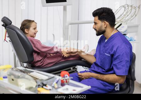 Un petit patient caucasien souriant sourit la main d'un jeune dentiste barbu après un traitement dentaire réussi et indolore à la clinique moderne Bright. Concept de stomatologie pédiatrique. Banque D'Images