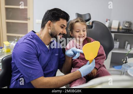 Bonne mignonne petite fille appréciant son beau sourire en train de regarder le miroir tandis que jeune barbu confiant dentiste le tenant dans la clinique dentaire moderne de bureau léger. Banque D'Images