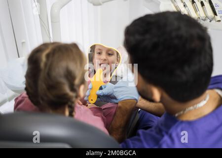 Bonne mignonne petite fille appréciant son beau sourire en train de regarder le miroir tandis que jeune barbu confiant dentiste le tenant dans la clinique dentaire moderne de bureau léger. Banque D'Images