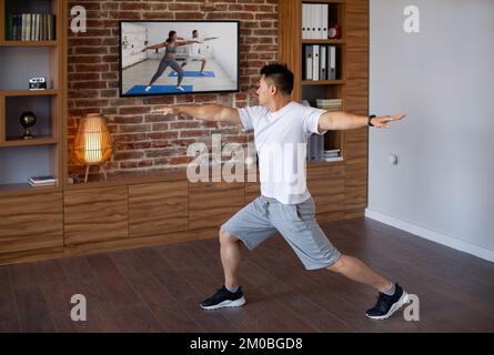 Concept d'entraînement domestique. Homme asiatique adulte actif faisant un étirement de fente avant, regardant un programme d'entraînement de fitness à la télévision Banque D'Images